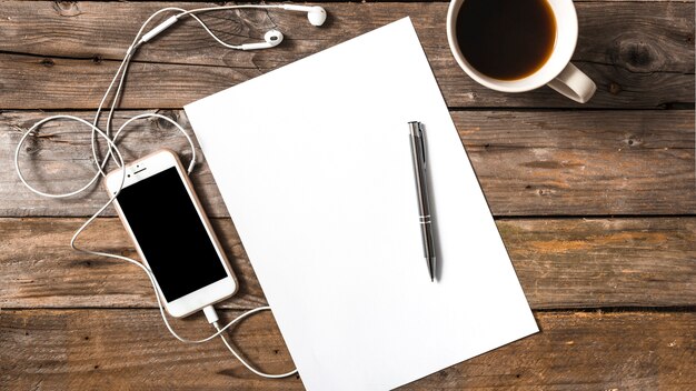 Мобильный телефон, подключенный к наушникам; ручка; бумага и чашка кофе на деревянном фоне