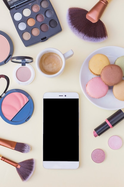 Cellphone screen; coffee with macaroons and cosmetics products on beige background