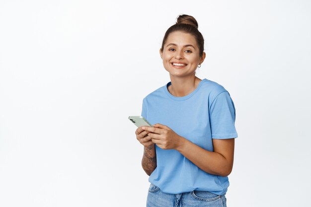 Концепция сотовых технологий. Улыбающаяся молодая женщина, использующая мобильный телефон на белом, в синей футболке