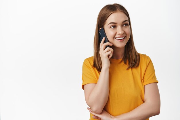 Cellular and technology concept. Young woman talking on mobile phone and smiling, wearing yellow t-shirt. Girl speaking with friend and looking aside, using smartphone, white background