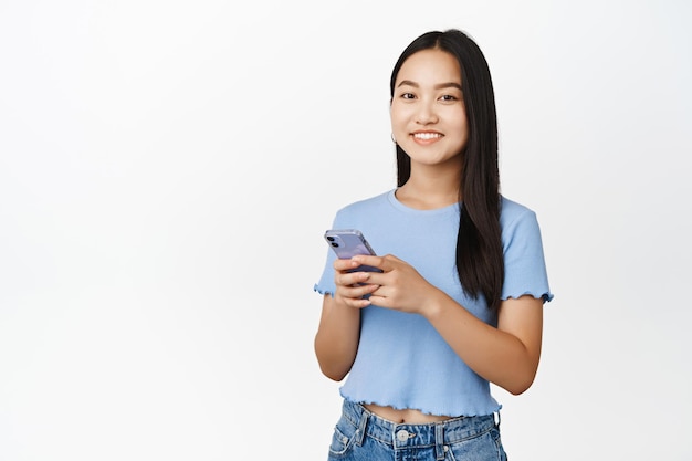 Cellular technology Smiling asian girl holding smartphone and looking happy at camera using mobile application white background