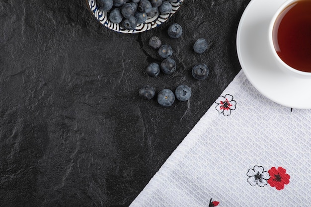 Ceramic bowl of delicious fresh blueberries and cup of tea on black surface