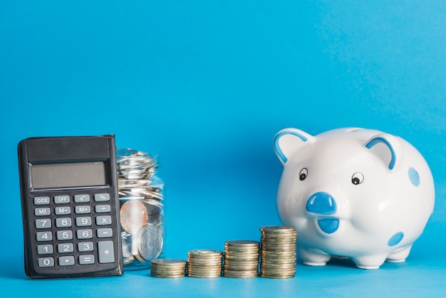 Ceramic piggybank; calculator; glass jar and stack of coins against blue background