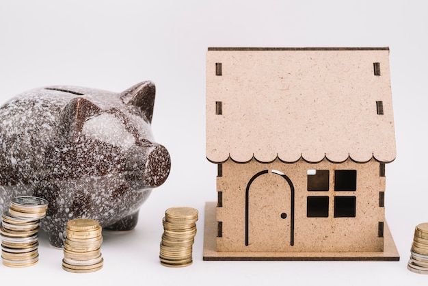 Ceramic piggybank with stack of coins near the cardboard house on white background