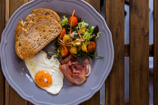 Ceramic plate of toast; fried egg; bacon and salad on ceramic plate over the wooden desk