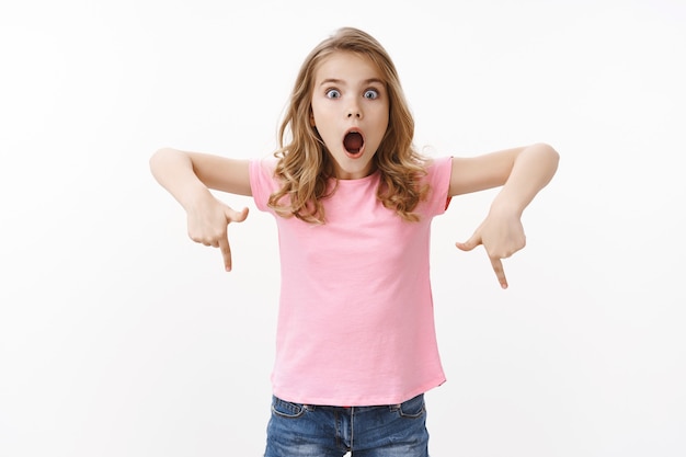 Charismatic amazed cute blond european girl in pink t-shirt, explain incredible thing, look astonished and excited, open mouth shouting impressed, pointing down, indicate bottom promo
