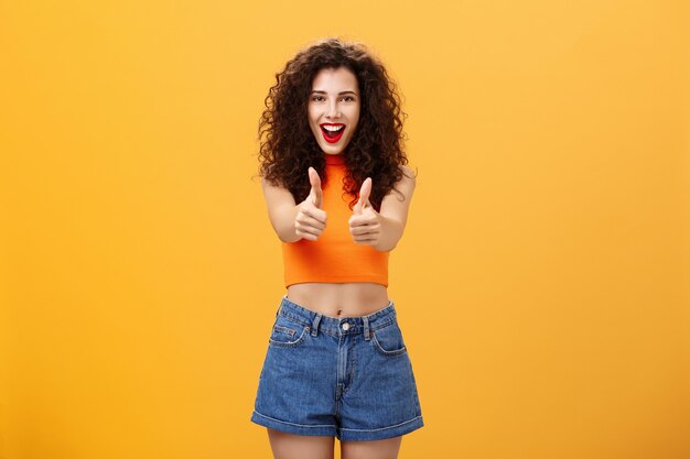 Charismatic ambitious and outgoing charming caucasian woman with curly hairstyle and red lipstick showing thumbs up gesture in like or approval smiling joyfully being supportive over orange wall.