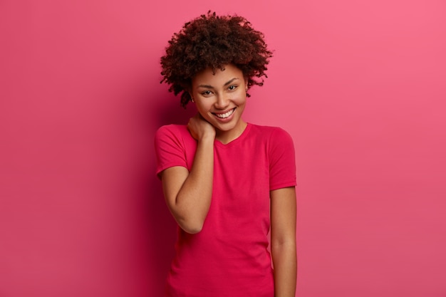 Харизматичная красивая кудрявая чувственная женщина трогает шею, счастливая улыбка на лице, любит проводить время с забавными людьми, носит повседневную футболку, позирует на розовой стене, имеет дружелюбный вид