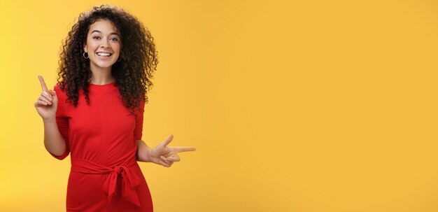 Charismatic cute and happy young curlyhaired european female in red dress dancing and pointing up an