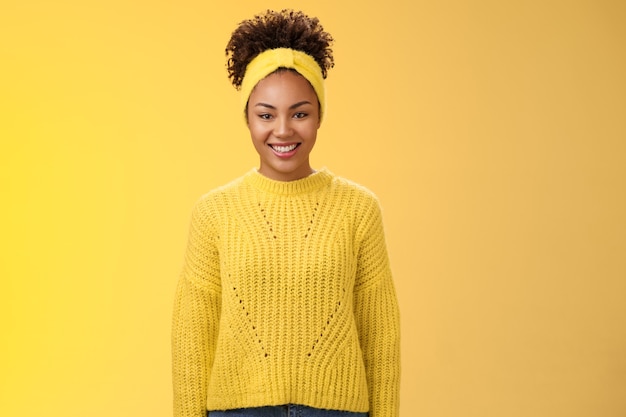 カリスマ的なフレンドリーな笑顔の黒人の女の子のヘッドバンドカーリーアフロヘアスタイルニヤリと笑って喜んで興奮しているユニバーティシーイベントに参加して、立ち上がる活気に満ちた黄色の背景を手伝ってください。