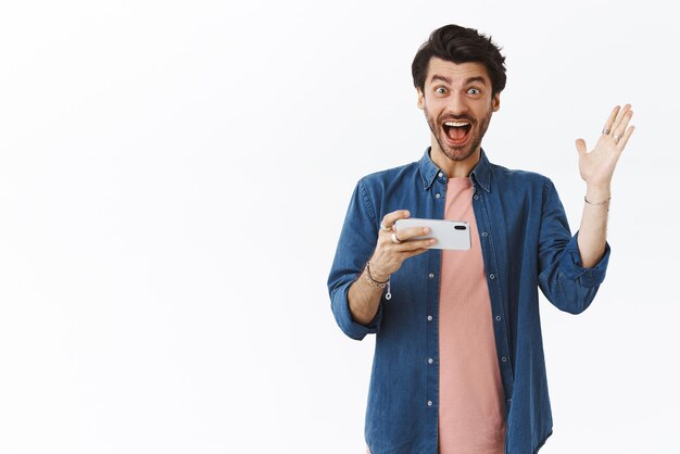 Charismatic happy 25s handsome man in casual outfit smiling and raising one hand in hooray yes gesture winning achieve goal passed level in favorite game holding smartphone white background