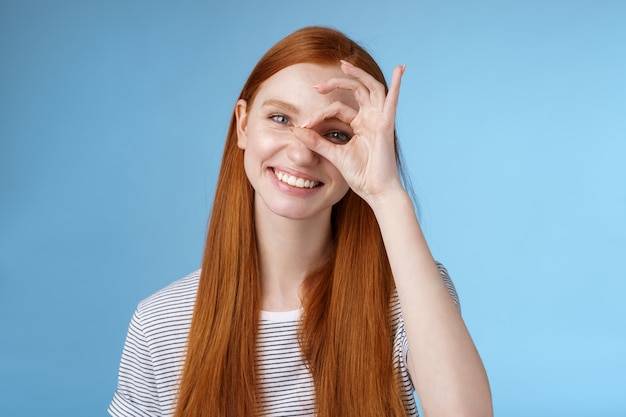 Charismatic happy adorable redhead teenage girl sincere eyes making circle eye show okay ok sign delighted like approve cool idea smiling satisfied achieve perfect score, standing blue background