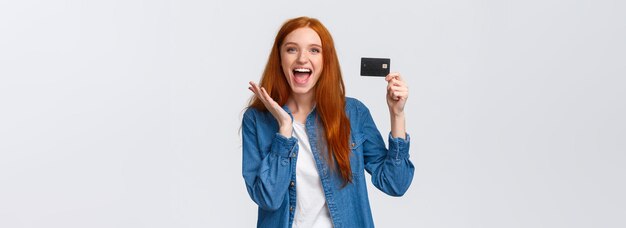 Харизматичная счастливая симпатичная рыжеволосая студентка в повседневной одежде с кредитной картой и лучом