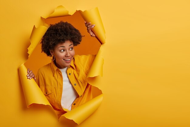 Charismatic joyful Afro woman with curly hairstyle turns away, looks on right side, wears fashionable shirt, stands in torn paper hole