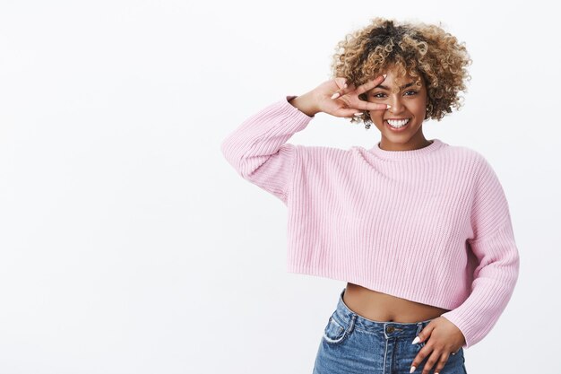 Charismatic optimistic and attractive african american woman in good mood expressing joy and delight showing victory or peace gesture on eye and smiling joyfully having fun over white wall