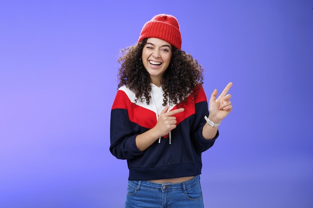 Charismatic playful young curly-haired european female winking joyfully at camera as pointing at upper right corner wearing hat and pullover as posing delighted over blue background, feeling warm.