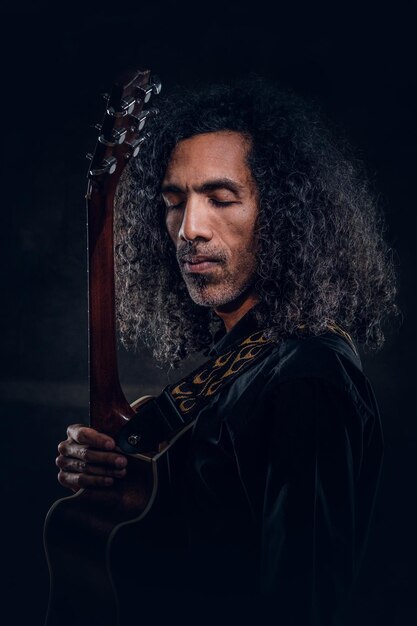 Charismatic rock singer is posing with his guitar at dark photo studio.