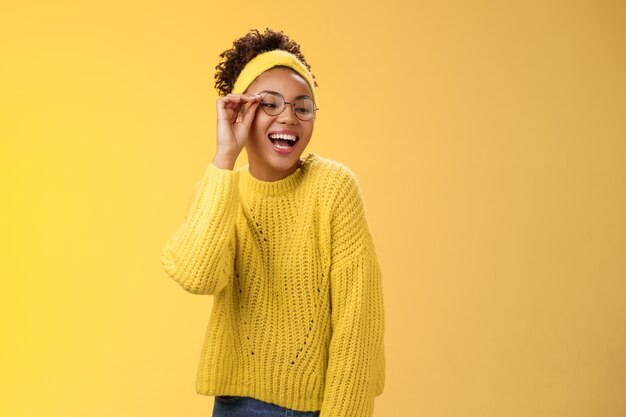 Charismatic stylish confident beautiful african-american joyful teenage girl touching glasses nose laughing broadly look down right corner grinning have fun joking enjoying, yellow background.