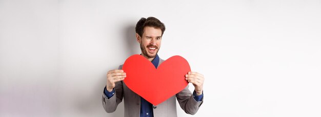 Charismatic young man winking and smiling showing big red heart cutout for valentines day date say l