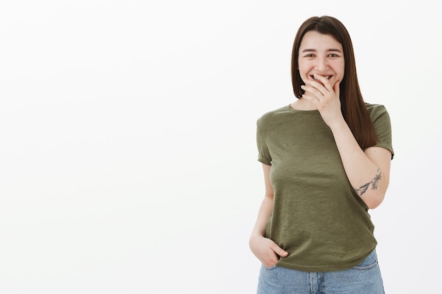 Charming 20s female brunette with tattoo on arm laughing and smiling covering mouth shy as giggling from hearing funny story, having fun talking amused and joyful over gray wall