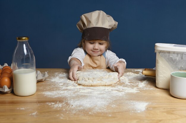 Charming 5 year old little girl wearing chef hat and apron kneading dough at kitchen counter