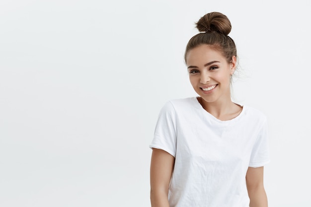 Charming adult caucasian woman with bun smiling broadly and glancing at camera, standing against white space