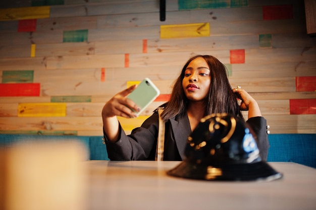 Charming african american woman model in black jacket relaxing in cafe during free time with mobile phone