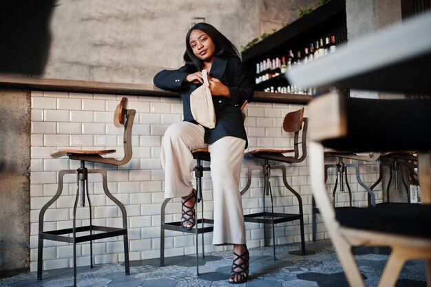 Charming african american woman model in black jacket and waist bag relaxing in cafe during free time