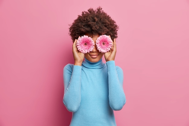 Charming African American woman with natural beauty, holds two gerberas on eyes, dressed in blue turtleneck, poses 