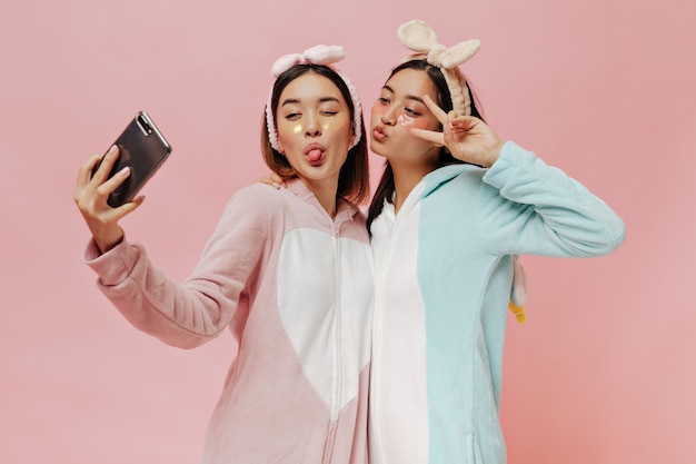 Charming Asian brunette women in soft pajamas and headbands make funny faces and take selfie on pink wall