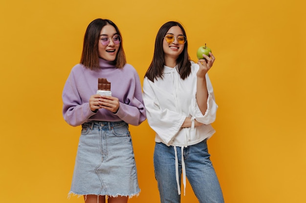 Charming Asian girl in denim skirt and purple sweater holds milk chocolate bar and looks at fruit of her friend Brunette woman in jeans and white hoodie hold fresh green apple