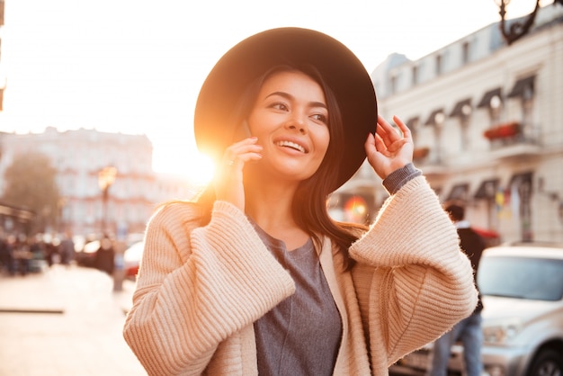 Charming asian woman adjusting her black hat while talking on mobile phone on city street