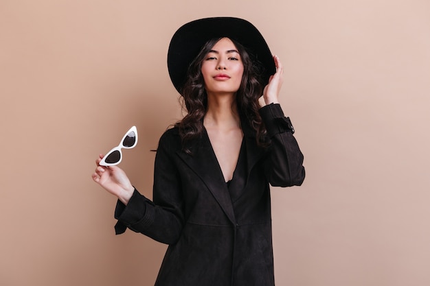 Charming asian woman in black coat looking at camera. Beautiful japanese woman in hat standing on beige background.