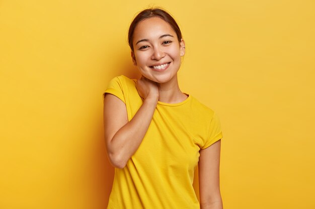 Charming Asian woman has cheerful expression, touches neck and tilts head, has pleasant talk with interlocutor, hears pleasant news, wears vivid casual t shirt, isolated on yellow wall