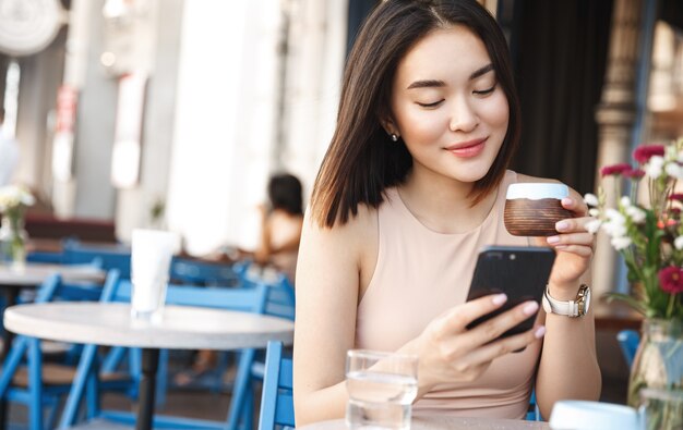 Charming asian woman with beautiful smile reading good news on mobile phone during rest in coffee shop