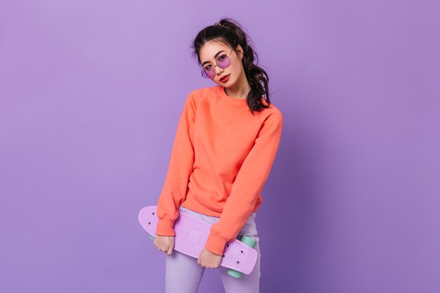 Charming asian woman with ponytail holding skateboard. Studio shot of adorable chinese young woman posing on purple background.