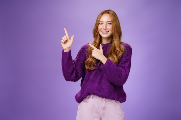 Charming assertive female shop assistant pointing at upper left corner to promote cool product smiling broadly feeling joyful and excited expressing friendly attitude as posing in purple sweater.