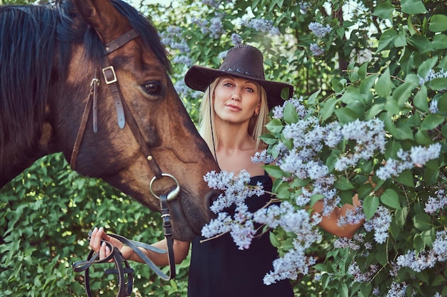 검은 옷을 입고 꽃밭에 말과 함께 서 있는 모자를 쓴 매력적인 아름다운 금발.