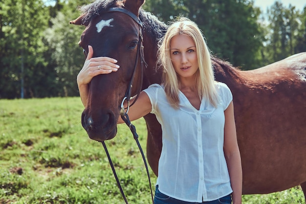 Bella bionda affascinante che indossa una camicetta bianca e jeans che abbraccia un cavallo in un campo verde.