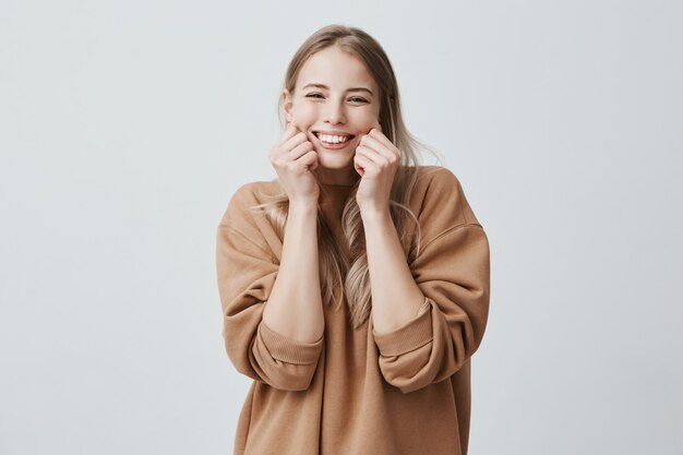 Charming beautiful female model smiling broadly wearing brown sweater, pinching her cheeks, mocking, having good mood and fun. Positive emotions and feelings