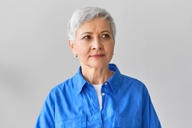 Charming beautiful middle aged gray haired woman on retirement posing isolated against blank wall  with copyspace for your text or advertising content, smiling thoughtfully