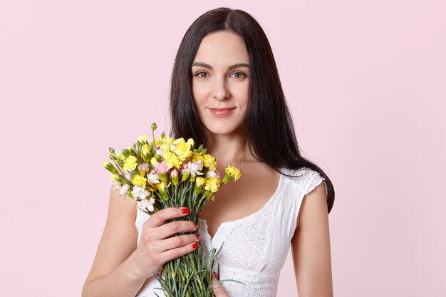Charming beautiful woman holds yellow pink flowers with one hand, looking directly at camera, feels pleased.