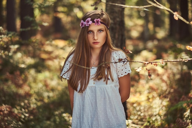 A charming beautiful woman in a white dress and purple wreath on head posing in a green autumn forest.