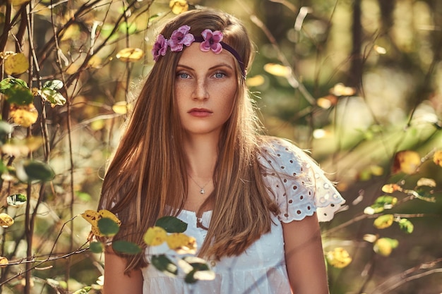 緑の秋の森でポーズをとる白いドレスと頭に紫色の花輪の魅力的な美しい女性。