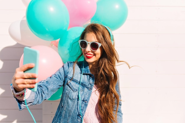 선글라스를 쓴 매력적인 생일 소녀는 스마트폰을 사용하여 화상 통화를 축하합니다. 헤드폰과 데님 재킷을 입은 멋진 젊은 여성이 파티에서 풍선으로 셀카를 만들고 있다