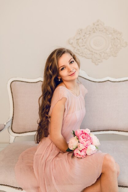 Charming blond girl sitting on a sofa. She has long curly hair and holds a bouqet of peonies in her hands.