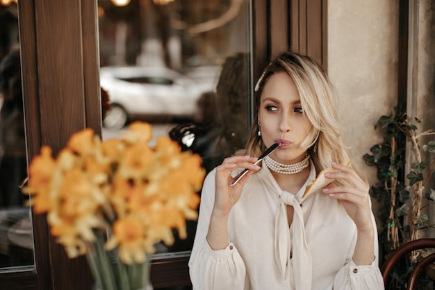 Charming blonde beautiful woman in white blouse and vintage pearl necklace sits in street cafe and licks knife