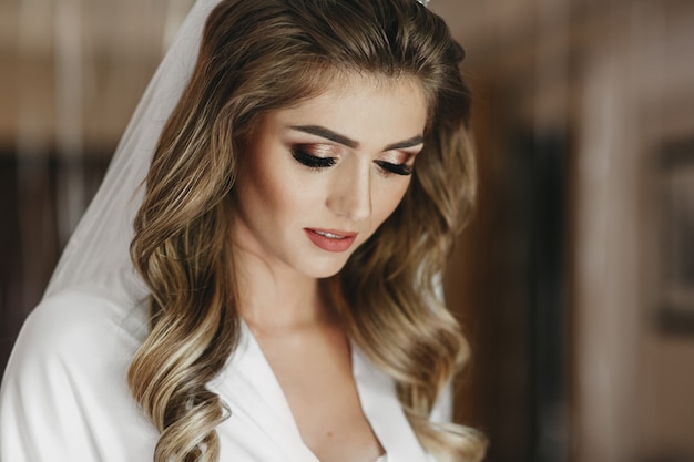 Charming blonde bride with curls and shiny skin poses in white silk robe in the room