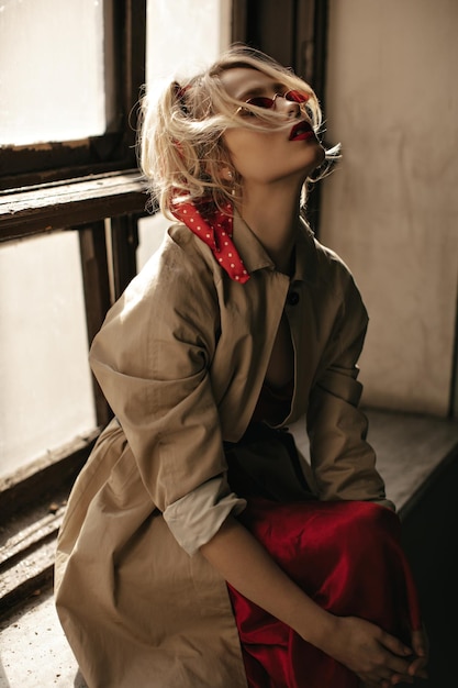 Charming blonde curly lady in red sunglasses beige trench coat and bright silk skirt looks up and sits on windowsill near window