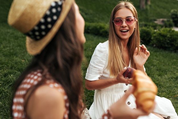 Charming blonde long-haired girl in red sunglasses and white dress holds apple, sits on rug on grass and talks to her brunette curly friend